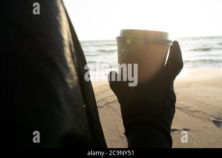 Coffee at the sea. Young woman with gloves holds a disposable cup of coffee on on a sunny morning at the beach. Stock Photo