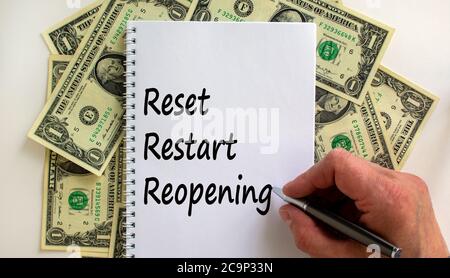 Male hand writing 'reset, restart, reopening' on white note, on white background. Dollar bills. Stock Photo