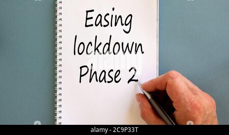 Male hand writing 'easing lockdown phase 2' on white note, isolated on blue background. Business concept. Stock Photo