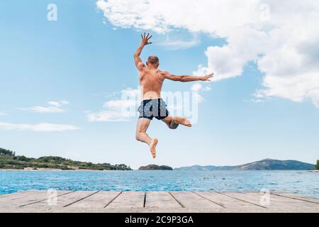 Middle-aged man dressed swimming trunks funny jumping to the waves from the boat pier as he having fun on merry vacation days on Adriatic sea coast. C