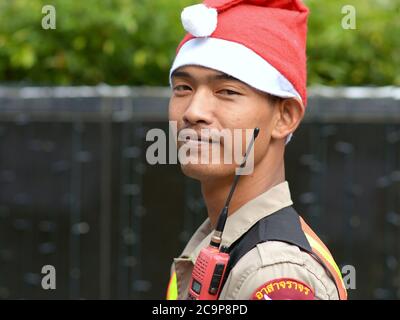 Uniformed Thai security guard with walkie-talkie (two-way radio) wears a Santa Claus hat and poses for the camera. Stock Photo