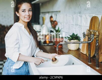 Young woman washes dishes with wooden brush with natural bristles at window in kitchen. Zero waste concept Stock Photo