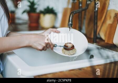 Young woman washes dishes with wooden brush with natural bristles at window in the kitchen. Zero waste concept Stock Photo