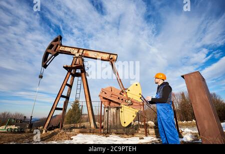 Petroleum engineer in work overalls and helmet holding clipboard, checking oil pumping unit, making notes. Man standing near oil pump jack. Concept of oil extraction and petroleum industry. Stock Photo