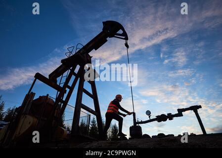 Silhouette of engineer in work vest and helmet working on petroleum pump jack. Oil worker using oil pump rocker-machine in oil field under evening sky. Concept of oil extraction and petroleum industry Stock Photo