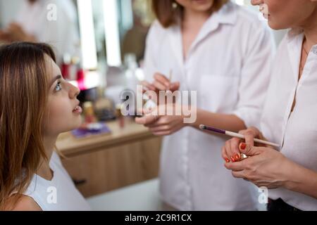 team of professional caucasian makeup artists working with young model. two make-up artist doing make-up in two hands and talk Stock Photo