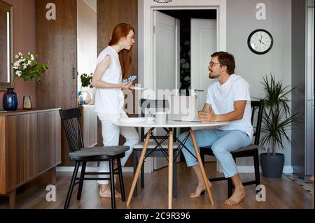 wife is outraged that her husband works a lot, caucasian married couple quarrelling in the kitchen. man sit with laptop Stock Photo