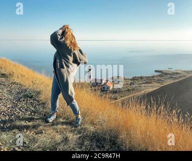 Side view of unrecognizable female standing on grassy hill and admiring amazing seascape during sunset and touching hair Stock Photo