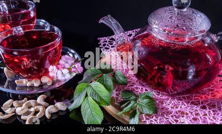 Green tea leaves or leaves of medicinal herbs lie on the table with a teapot and mugs of red tea, cashew nuts and clove flowers. Tea parties, tea trad Stock Photo