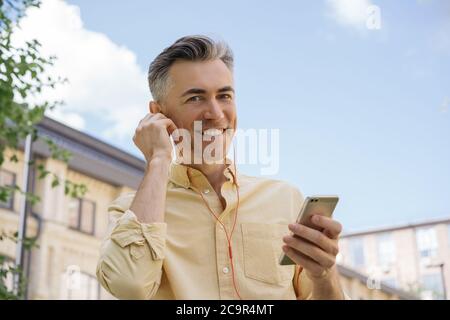 Handsome mature man holding mobile phone, listening to music in red earphones, looking at camera, smiling Stock Photo