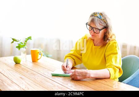 Calm elderly female in bright clothes and eyeglasses sitting at wooden table at home and writing in notebook Stock Photo
