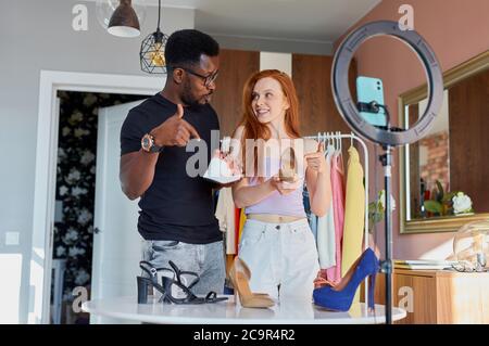 two fashion bloggers talk at camera at home, young diverse man and woman broadcast on camera together, give useful tips on combining things Stock Photo