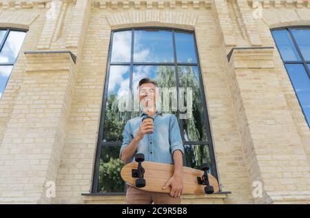 Happy skater holding longboard, drinking coffee, walking on the street, looking at camera and smiling Stock Photo