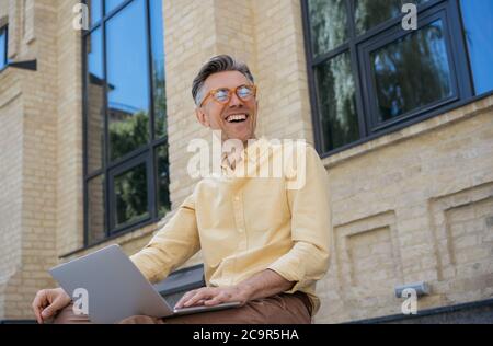 Happy stylish businessman using laptop computer, typing on keyboard, laughing. Handsome mature man working project, planning start up Stock Photo