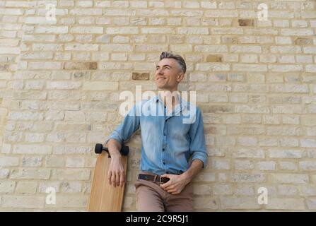 Stylish mature fashion model wearing casual clothes, posing for pictures. Happy skater with longboard, standing near wall Stock Photo