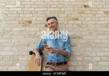 Happy mature skater with longboard, standing near wall, holding smartphone, smiling. Portrait of modern middle aged businessman using mobile phone Stock Photo