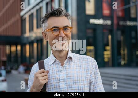 Portrait of handsome mature businessman wearing stylish eyeglasses, walking on street, looking at camera, smiling. Successful business and career Stock Photo