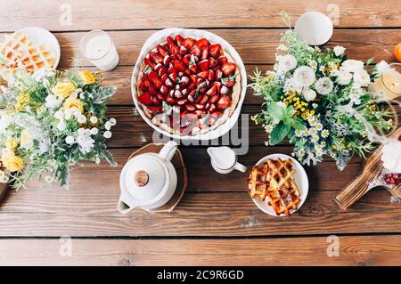Top view of delicious strawberry cake and homemade waffles arranged on wooden table with flower bouquets in countryside on sunny day Stock Photo