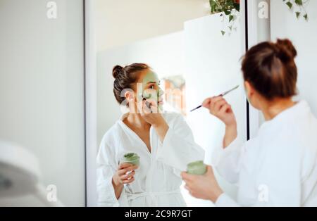 Young brunette female in white bathrobe smearing mud mask on face while standing in front of mirror during spa procedure at home Stock Photo