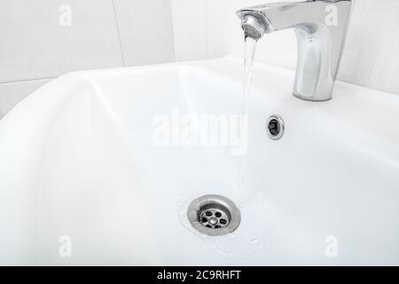 open chrome faucet water flow in the ceramic wash basin in the bathroom with a beige tile on wall. Stock Photo