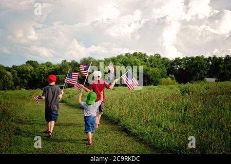 Three boys holding American Flags while walking on a country path in a field of green waving the flags Stock Photo