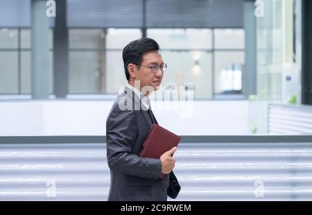 Chinese businessman smiling and walking with his hand holding a diary. Modern layout setting. Stock Photo