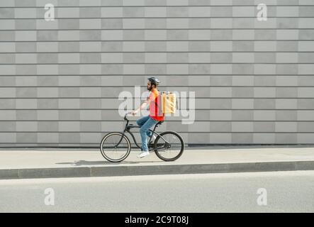 Delivery man on bicycle. Young guy with beard and big backpack rides along path in city