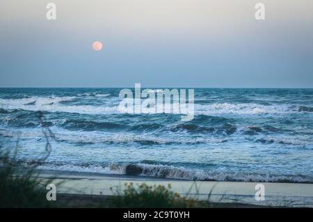Rise of full moon over the sea at sandy beach and rough waves. Southern France. Stock Photo