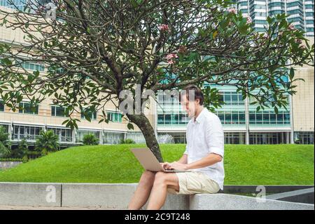 Man businessman or student in casual dress using laptop in a tropical park on the background of skyscrapers. Mobile Office concept Stock Photo