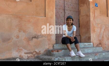 Woman sitting on steps in front of doorway with cap and sneakers in Bologna Italy looking at photographer Stock Photo