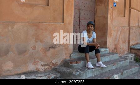 Woman sitting on steps in front of doorway with cap and sneakers in Bologna Italy Stock Photo