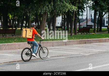 Courier bicycle delivery service. Man with large yellow backpack ride along road in park