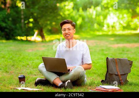 Smiling asian student guy sitting with laptop outdoors, preparing for exams Stock Photo