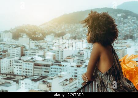 Side view of thoughtful sleepy an African-American girl leaning against a balcony fence and pensively observing typical hilly Brazilian cityscape Stock Photo