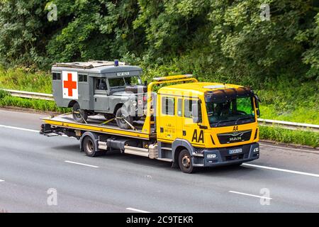 AA Van recovery truck carrying 1997 Land Rover Military Army ambulance  Side view of AA rescue breakdown recovery MAN double cab roadside recovery lorry truck transporter,  transporting vehicle driving along  M6, Chorley UK; Vehicular traffic, transport, modern on the 3 lane highway. Stock Photo