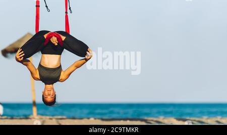 young woman doing yoga exercises with swing upside down on the beach in front of the sea on a sunny day Stock Photo