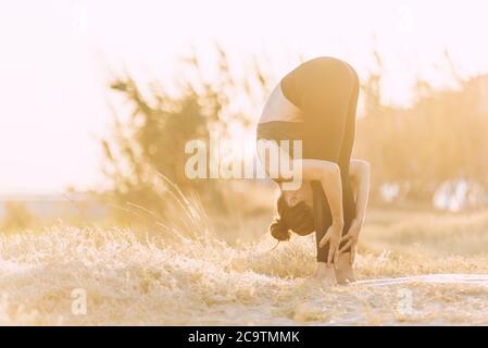 young woman doing stretching yoga exercise at golden sunset outdoors next to greenery Stock Photo