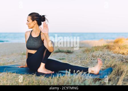 brunette young woman doing yoga exercise on the ground outdoors by the sea at sunset Stock Photo