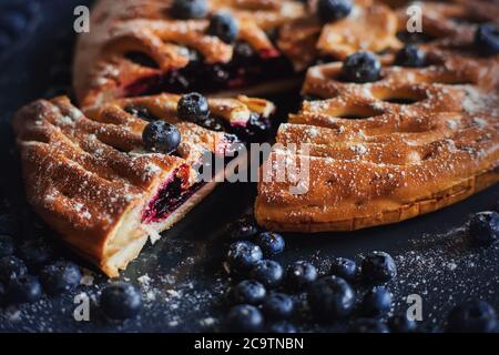 Homemade sweet delicious pie with blueberry jam, a piece of which is cut off and lies on a baking sheet among ripe blueberries. Homemade cake. Stock Photo