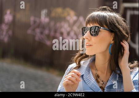 Outdoor portrait of yong beautiful happy smiling woman wearing stylish sunglasses, black polka dot blouse, blue mom jeans, with small quilted bag. Mod Stock Photo