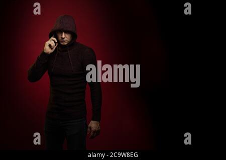 A man in a jumper with a hood and with a phone in his hands poses against a dark red background Stock Photo
