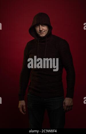 A man in a jumper with a hood and with a phone in his hands poses against a dark red background Stock Photo