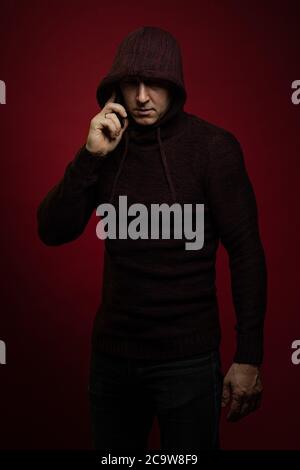 A man in a jumper with a hood and with a phone in his hands poses against a dark red background Stock Photo