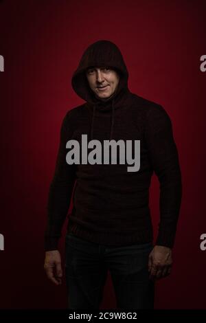 A man in a jumper with a hood and with a phone in his hands poses against a dark red background Stock Photo