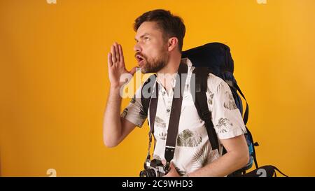 Young man traveler with backpack speaking loud with hands on the side of his face isolated on the orange background. High quality photo Stock Photo