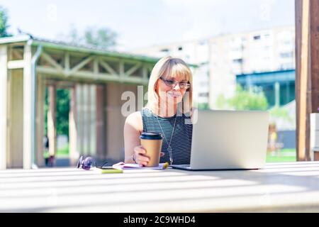 Adult Woman in glasses having video call with laptop outside in park. Happy and smiling senior working and drinking coffee. Using computer. Distance Stock Photo