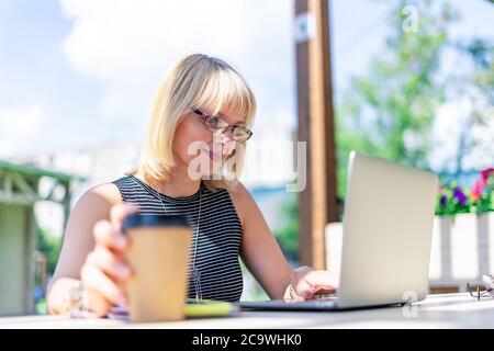 Adult Woman in glasses having video call with laptop outside in park. Happy and smiling senior working and drinking coffee. Using computer. Distance Stock Photo