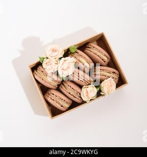 Flat lay of many chocolate french dessert macarons in craft box with beautiful beige roses on the white background. Stock Photo