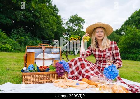 Girl in red checkered dress and hat sitting on white knit picnic blanket reading book and drinking wine. Summer picnic on sunny day with bread, fruit, bouquet hydrangea flowers. Selective focus Stock Photo