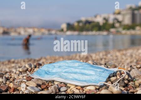 Discarded used face mask lies on a pebble beach, beachgoers are relaxing by the sea in the background. Stock Photo